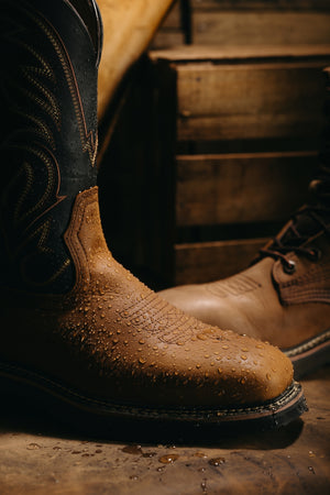 A close-up of a brown leather cowboy boot with water droplets on the vamp, standing on a rustic wooden surface with another boot and wooden crates in the background.